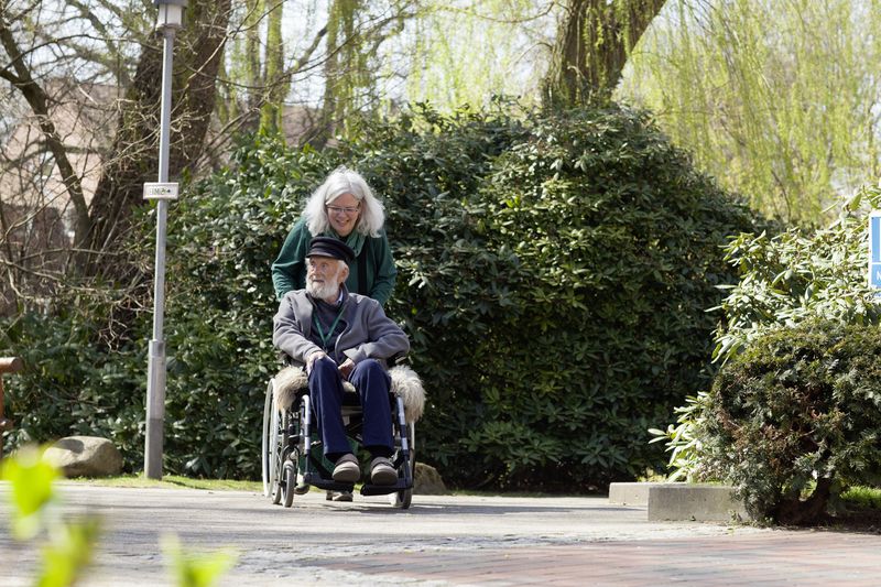 Mittelalte Frau mit langen grauen Haaren schiebt alten Mann mit weißem Bart im Rollstuhl durch den Park.
